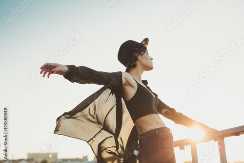 Flying like a beautiful bird in the wind teenager girl. Black clothes and hat. Relaxed dreaming mood. Summer evening in big city photo