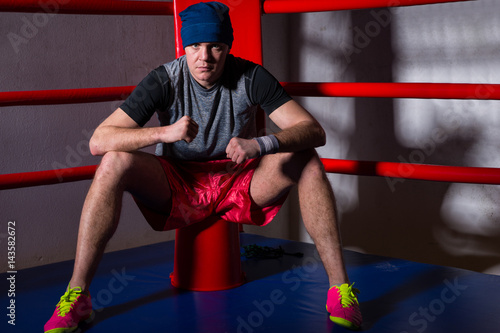 Athletic male boxer sitting near red corner of a regular boxing ring