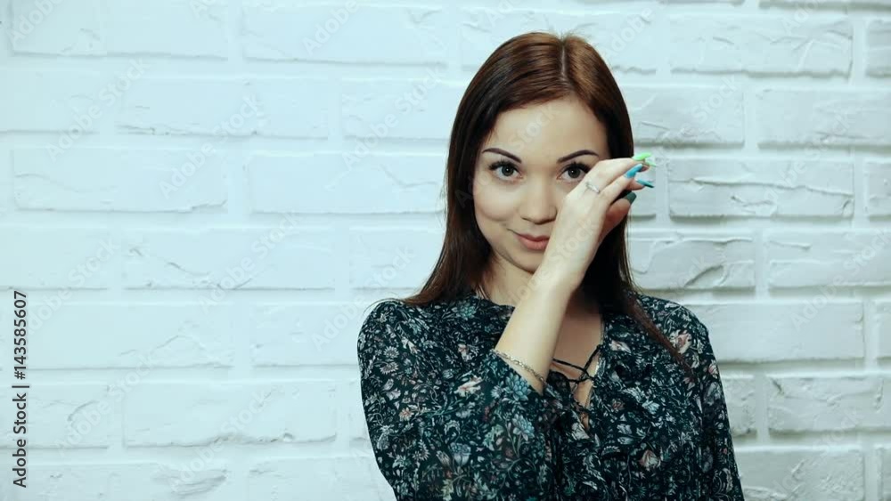 A young brown-haired woman adjusts her eyelashes, posing, looking at the camera on a white brick wall background. A young brown-haired woman adjusts her eyelashes, posing, looking at the camera on a white brick wall background.