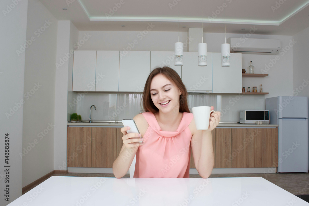 Beautiful girl in home using smartphone, drinking tea and smiling in kitchen Beautiful girl in home using smartphone, drinking tea and smiling in kitchen