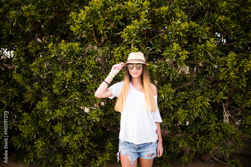 Woman smiling while leaning on wall outdoors ,happy stylish woman in glasses and hat standing at green leaves wall, summer travel concept. © F8  \ Suport Ukraine
