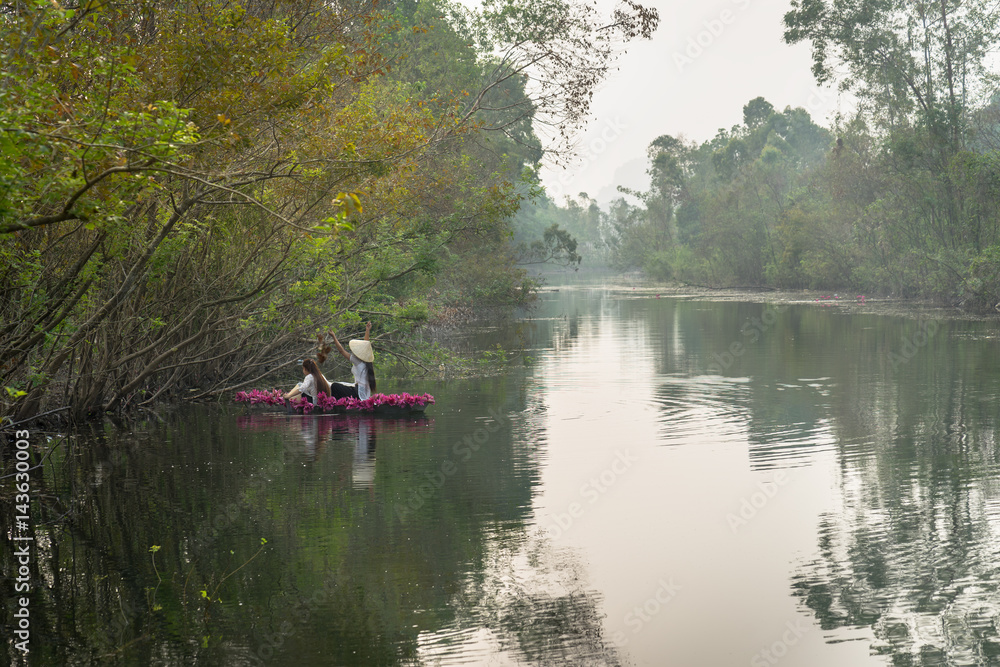 River scene with a boat carrying girls wearing traditional dress Ao Dai, conical hat, and flower.