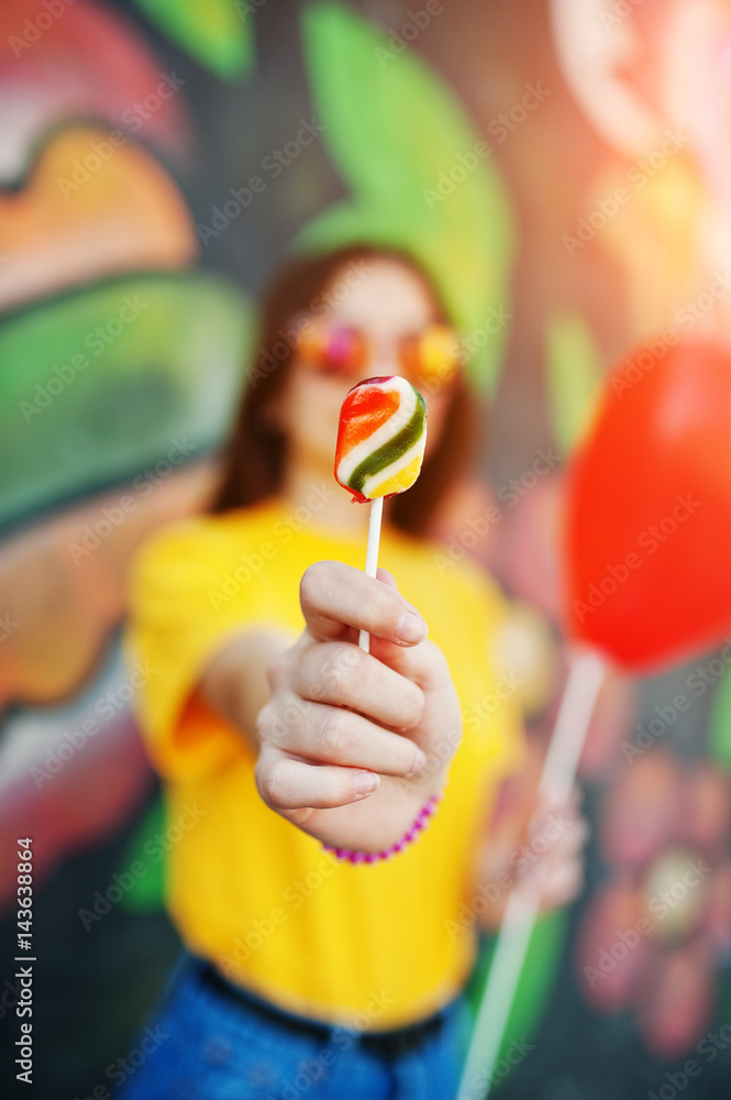 Beautiful teenage showing lollipop, wear on glasses, heart balloon at hand, yellow t-shirt near graffiti wall. Beautiful teenage showing lollipop, wear on glasses, heart balloon at hand, yellow t-shirt near graffiti wall.