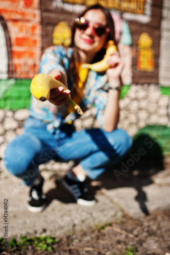 Beautiful fun teenage girl speaking bananas like phone, wear yellow t-shirt, jeans and sunglasses near graffiti wall.