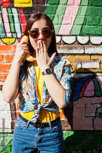 Beautiful fun teenage girl speaking bananas like phone, wear yellow t-shirt, jeans and sunglasses near graffiti wall.