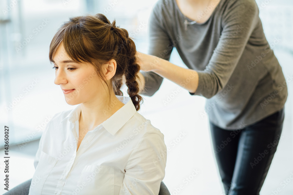 Fototapeta premium Super styling. Close up of a hairdresser braiding her clients hair in trendy weave plait while sitting in hairdressing salon. European model. Beauty lifestyle concept.