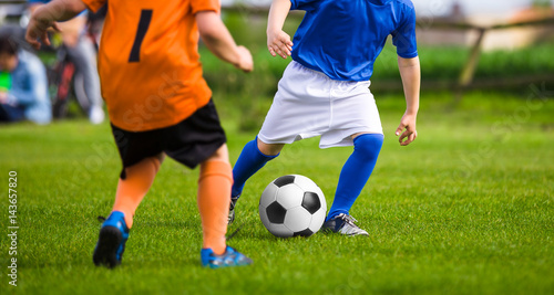 Two young boys in orange and blue jersey shirts kicking soccer match on the green grass field. Summertime sport soccer activity for kids. Children running and competing for the ball.