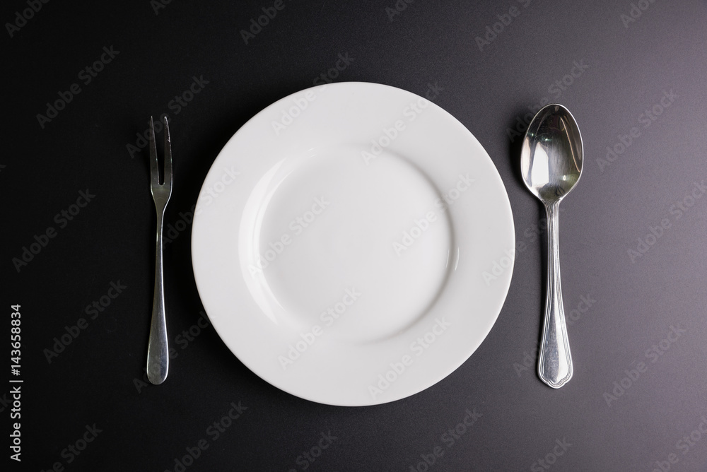 White dish, Spoon and fork isolated on black background with copy space image for your text. White dish, Spoon and fork isolated on black background with copy space image for your text.