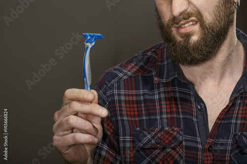 No shave. Cropped studio shot of a disgusted bearded guy holding a razor on dark background.