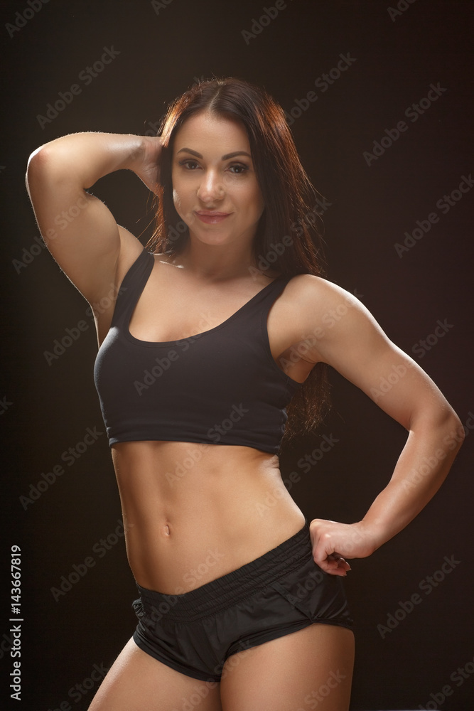 Perfect stunner. Portrait of a charming muscular woman posing at the studio fixing her hair looking to the camera on black background. Perfect stunner. Portrait of a charming muscular woman posing at the studio fixing her hair looking to the camera on black background.