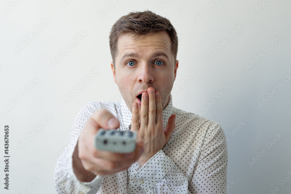 Naklejka premium A surprised young man switches channels by clicking on the buttons. Shock from the information on TV.