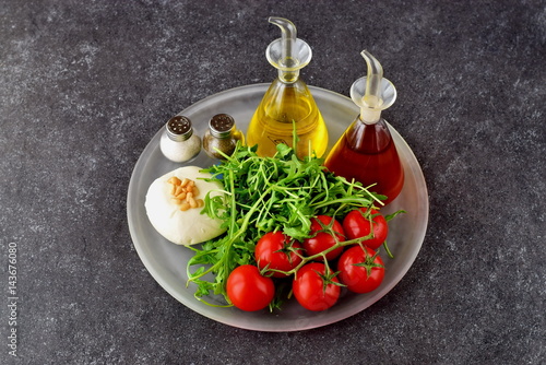 Ingredients for the traditional Italian caprese salad on a glass trey on a grey background with bottles of olive oil and wine vinegar.