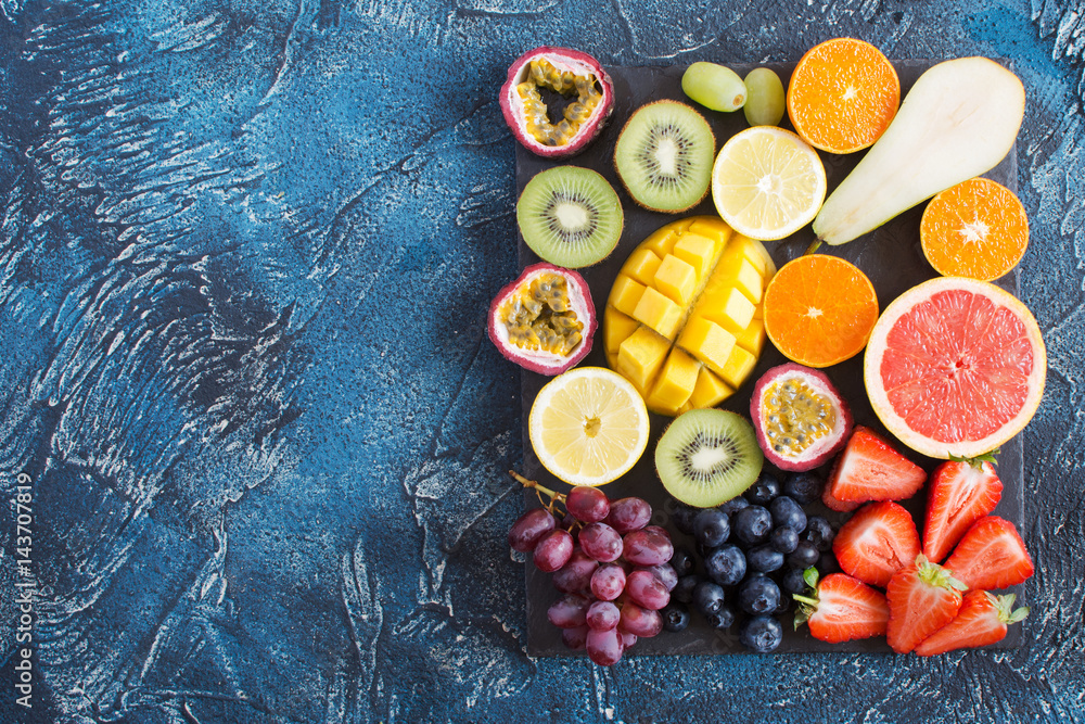 Top view of different raw fruits platter: oranges, lemons, kiwis, grapefruits, strawberries, grapes, blueberries, pears, passion fruit, mango on the black slate on the dark blue background Top view of different raw fruits platter: oranges, lemons, kiwis, grapefruits, strawberries, grapes, blueberries, pears, passion fruit, mango on the black slate on the dark blue background