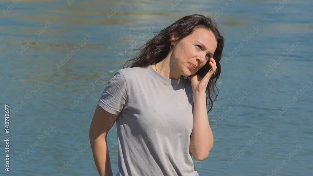Girl with mobile phone by the water. A woman is talking on the phone on the river embankment on a sunny day. Talk on the phone in the fresh air. Girl with curly hair in gray clothes. Girl with mobile phone by the water. A woman is talking on the phone on the river embankment on a sunny day. Talk on the phone in the fresh air. Girl with curly hair in gray clothes.