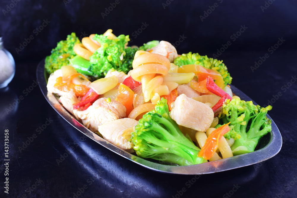 Stir fried chicken fillet with vegetables and pasta on a metal tray on a black abstract background.
