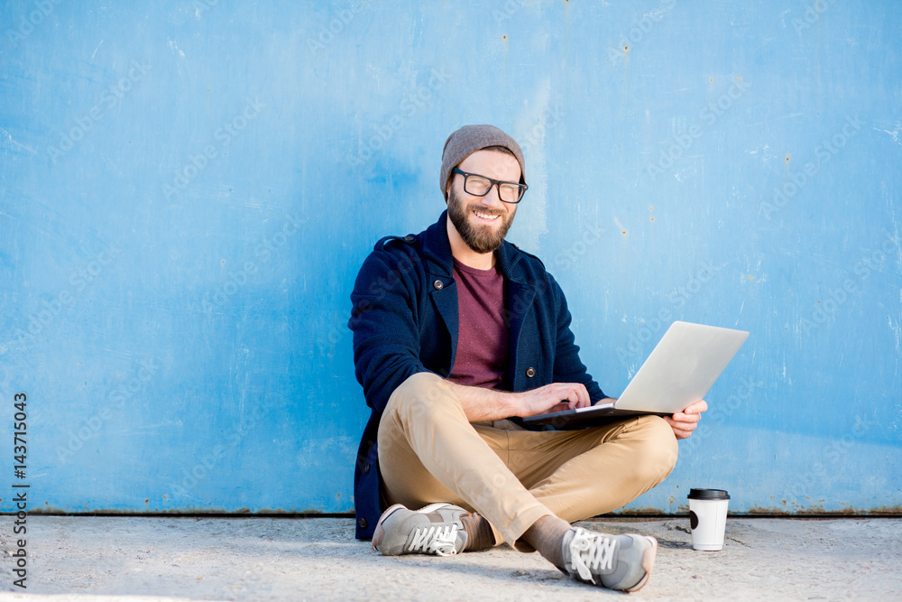 Stylish man dressed casual in sweater and hat working with laptop sitting near the blue wall background Stylish man dressed casual in sweater and hat working with laptop sitting near the blue wall background