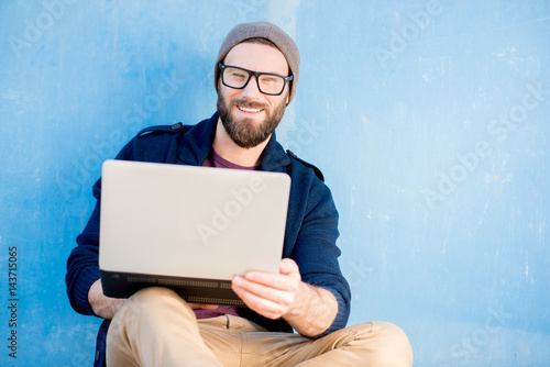 Stylish man dressed casual in sweater and hat working with laptop sitting near the blue wall background photo