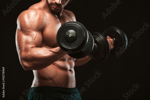 Handsome power athletic man in training pumping up muscles with dumbbells in a gym. Fitness muscular body isolated on dark background.