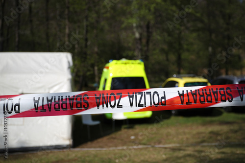 Red and white police line around a crime scene tent in a forest with ambulance,police and coroner vehicles