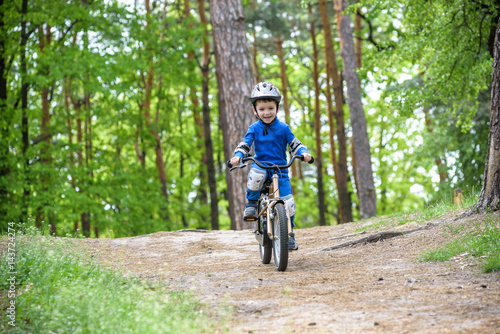Happy funny little kid boy in colorful raincoat riding his first bike on cold day in forest. Active leisure for children outdoors. Happy carefree childhood concept.