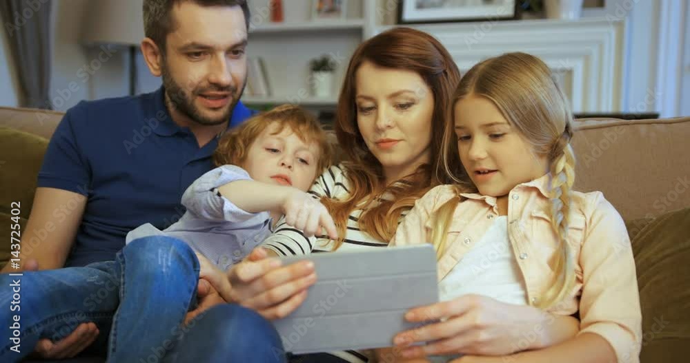 Mother, father, son and daughter using tablet, watching movie. Family spending time together at home. Happy family Mother, father, son and daughter using tablet, watching movie. Family spending time together at home. Happy family