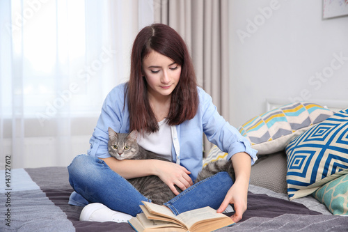 Beautiful young woman with cute cat reading book while sitting on bed at home