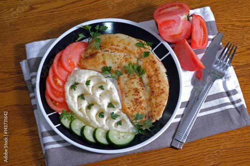 Fried tilapia fish fillets on plate with mashed potatoes, cucumber, tomatoes, parsley. 
