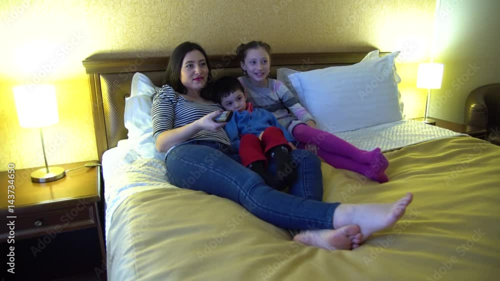 A young mother with two children, lying in hotel in bed and watch TV. Use the TV remote control. A young mother with two children, lying in hotel in bed and watch TV. Use the TV remote control.