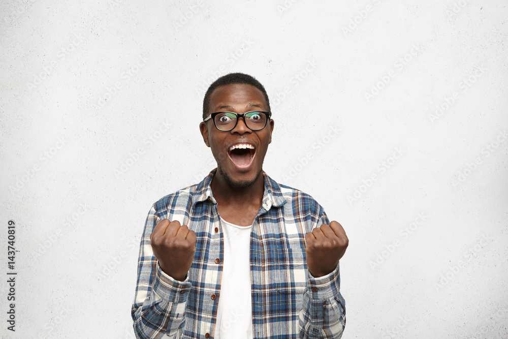 Omg! Portrait of shocked and amazed young African man in stylish clothing clenching fists and screaming in excitement, looking at camera in full disbelief after his favorite football team won match Omg! Portrait of shocked and amazed young African man in stylish clothing clenching fists and screaming in excitement, looking at camera in full disbelief after his favorite football team won match