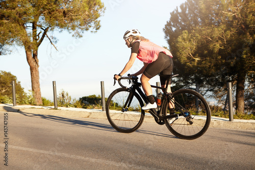 Beautiful athletic girl riding racing bike uphill on road against nature background. Young European blonde woman wearing white helmet, sunglasses, black-pink sportswear and sneakers cycling outdoors