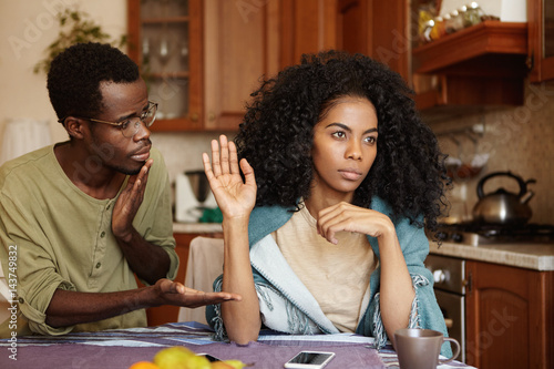 Regretful guilty young Afro-American man in glasses offering hand to his angry girlfriend as a sign of reconciliation after serious quarrel but woman seems refusing all apologies and excuses