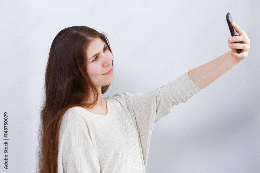Portrait of a young woman holding a smartphone in her outstretched hand and photographing herself. Making selfie. Grey background. Portrait of a young woman holding a smartphone in her outstretched hand and photographing herself. Making selfie. Grey background.