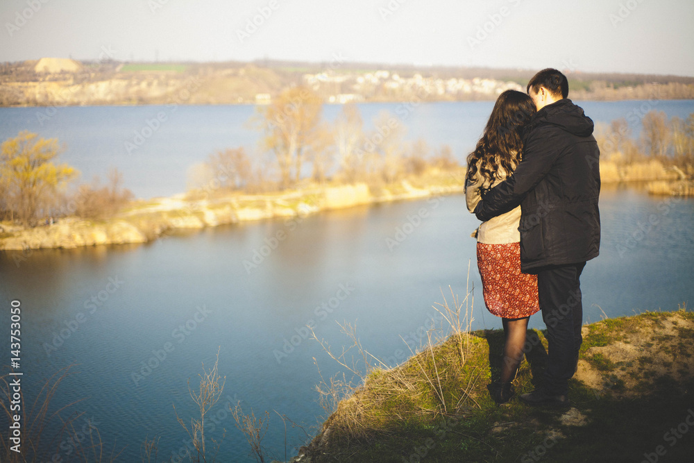 Husband and wife on the shore of the lake with rocky shores, early spring. Silhouettes of lovers who go into the water on the background of sunset over a lake.
