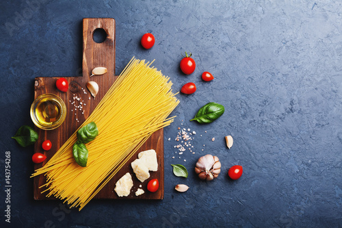Italian food background with uncooked spaghetti, tomato, basil leaves, cheese, garlic and olive oil on blue stone table top view in flat lay style. Ingredients for cooking pasta with space for recipe.