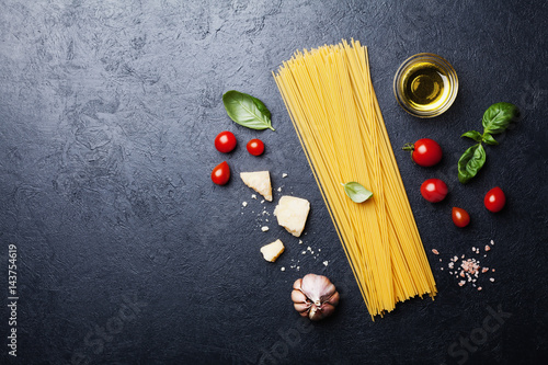 Italian food background with uncooked spaghetti, tomato, basil leaves, cheese, garlic and olive oil on black table top view in flat lay style. Ingredients for cooking pasta with space for recipe.