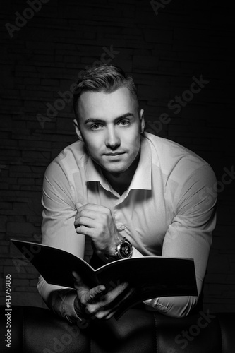 Young handsome businessman sitting in confident pose on leather couch and thinking. With magazine in hand