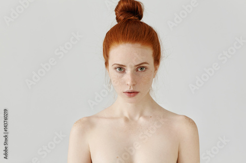 Isolated portrait of beautiful young Caucasian redhead woman with hair bun and perfect clean skin with freckles standing at grey studio wall with shoulders naked, having serious facial expression photo