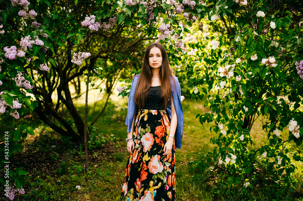 Happy and attractive beautiful artistic young model girl in colorful dress with long hair in summer lilac garden with bushes and flowers posing for camera. Spring concept. Happy and attractive beautiful artistic young model girl in colorful dress with long hair in summer lilac garden with bushes and flowers posing for camera. Spring concept.