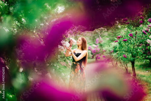 Portrait of an attractive beautiful young model girl in colorful dress with long hair in summer lilac garden with bushes and flowers. Spring concept. Photo through the brunches.