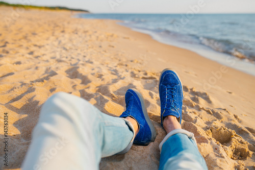 Legs of a man in light blue jeans and blue shoes laying on a sand on beach beside the sea