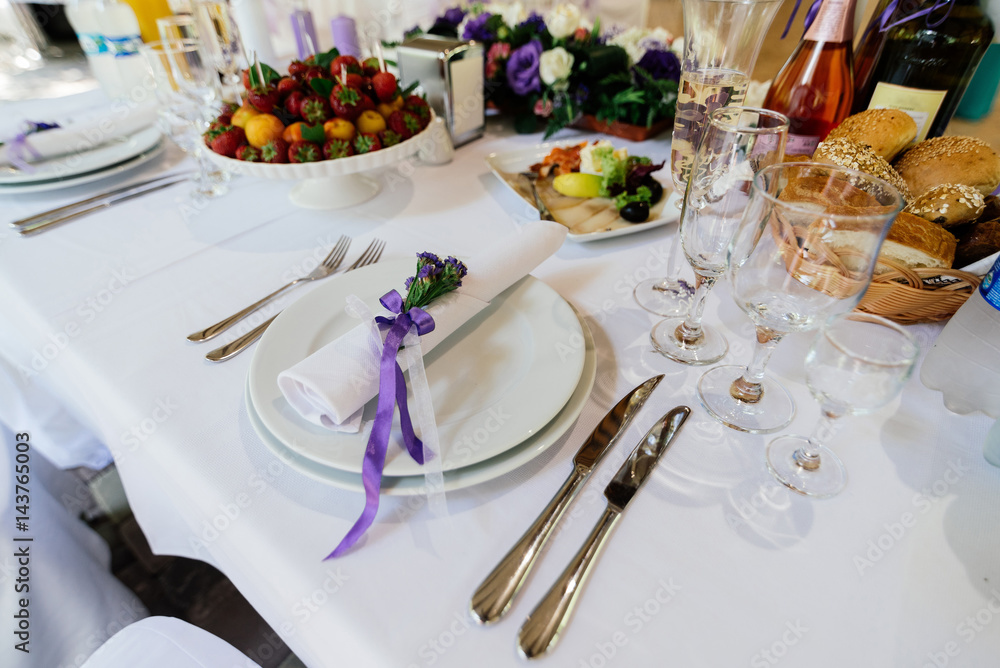 Fototapeta premium White napkin with purple flowers on a white empty plate on a dining table. Table setting. Table served for edding dinner, close up view