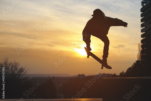 Obraz na plátně The teenager in a sweatshirt and a cap jumps with a board in the city against the backdrop of the urban sunset