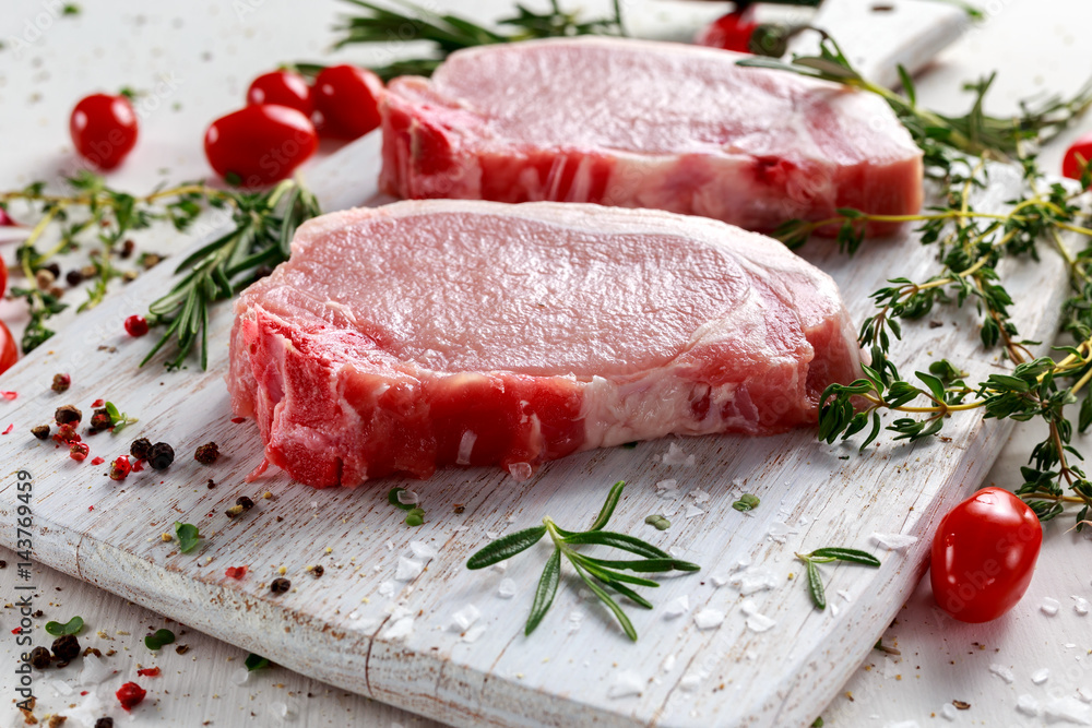 Raw Pork Loin chops on a cutting board with herbs, rosemary, thyme, chilli, salt, pepper on white cutting board.