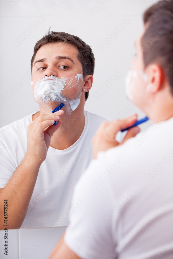 photo of handsome young man shaving and looking at himself in the mirror