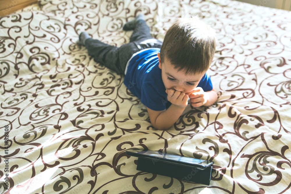 Little boy looking at tablet while lying on bed, natural light. Little boy looking at tablet while lying on bed, natural light.