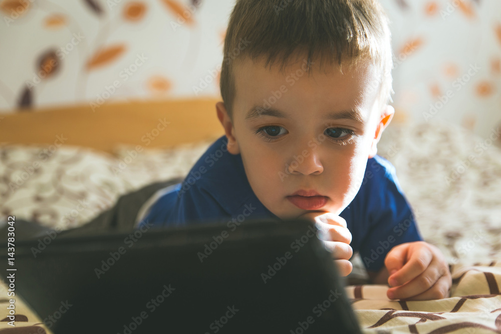 Little boy looking at tablet while lying on bed, natural light. Little boy looking at tablet while lying on bed, natural light.