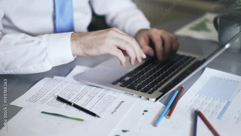 Close up of a man wearing a blue tie writing down numbers and typing at his laptop keyboard. Locked down real time close up shot Close up of a man wearing a blue tie writing down numbers and typing at his laptop keyboard. Locked down real time close up shot