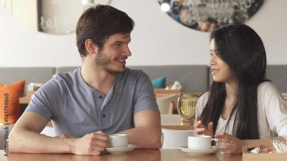 Two young friends of different genders chatting at the cafe. Handsome caucasian guy and pretty asian girl looking at each other. Attractive brunette woman touching her hair Two young friends of different genders chatting at the cafe. Handsome caucasian guy and pretty asian girl looking at each other. Attractive brunette woman touching her hair