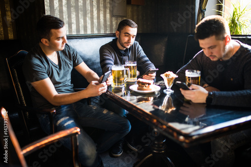 Male friends sitting in smartphones drinking beer at bar or pub. Smartphones social network dependence.