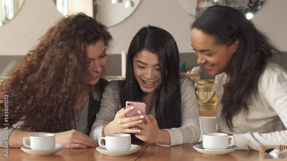 Three cute girls watching something funny on smartphone at the cafe. Pretty brunette lady holding cellphone in her hands. Attractive african american and caucasian women sitting from the sides of Three cute girls watching something funny on smartphone at the cafe. Pretty brunette lady holding cellphone in her hands. Attractive african american and caucasian women sitting from the sides of